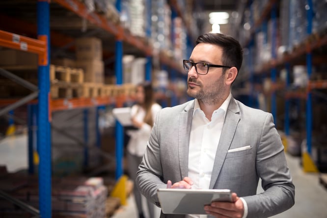 Man wearing a suit and glasses on warehouse floor holding a tablet.
