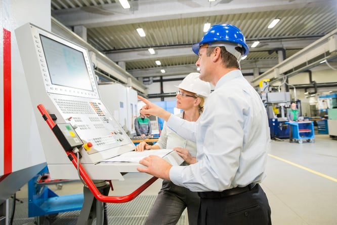 Man and women wearing white and blue hard hats in a warehouse looking at a machine screen. 