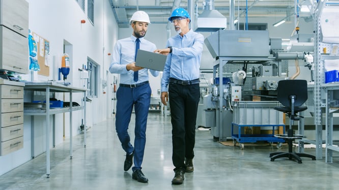 Middle aged man and a younger man wearing hard hats in a manufacturing warehouse talking and holding a laptop.
