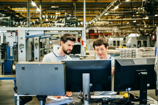 Tow men on a plant floor troubleshooting in front of three monitors